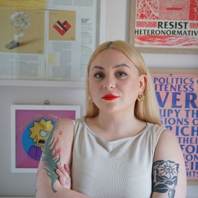 a blonde woman with tattoos sitting in front of a wall of colourful posters