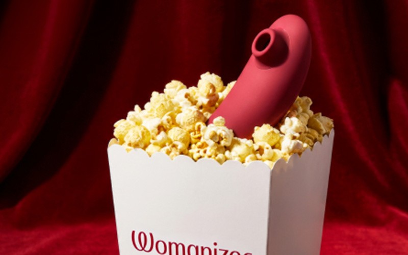 A red background with a Womanizer popcorn box, filled with popcorn and a Womanizer toy poking out of the box.