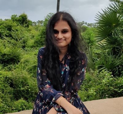 a woman with brown hair and a floral dress sits smiles in front of leafy green foliage